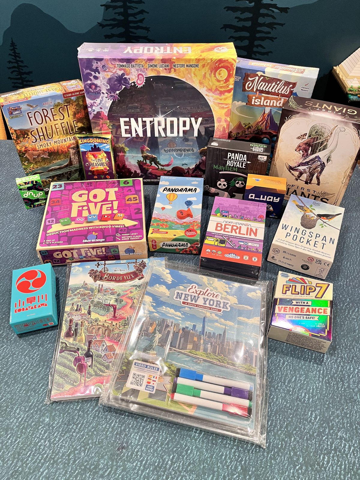 A Gen Con haul of board game boxes stacked on a table, including Entropy, Forest Shuffle: Smoke Mountain, Nautilus Island, Got Five!, Wingspan Pocket, Berlin, and Flip 7.