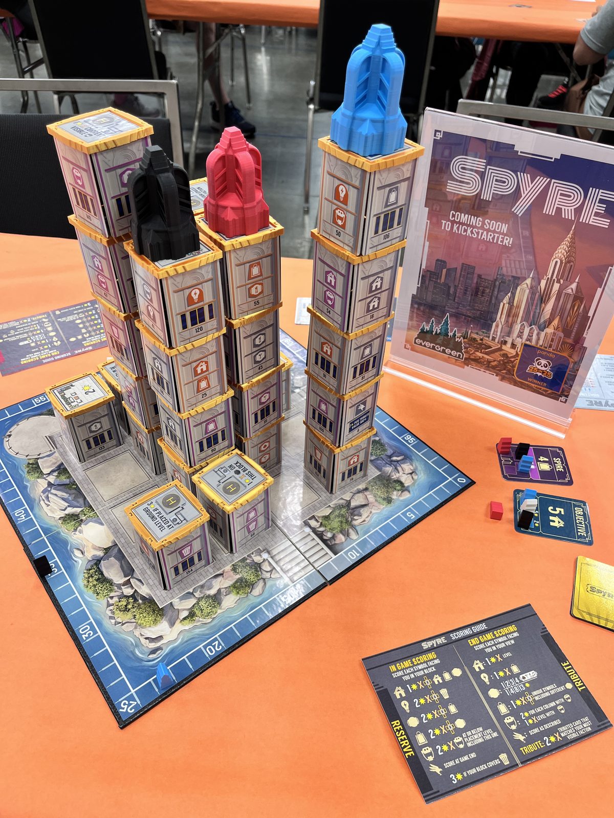 A finished game of Spyre, with several towers built up in black, red, and blue, beside a Spyre standee reading Coming Soon to Kickstarter.